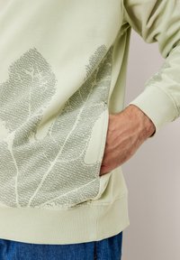 Felpa verde chiaro con un design a motivi di foglie in verde scuro, con polsini a coste e una tasca frontale visibile.