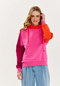 Růžová a oranžová barevně blokovaná mikina s kapucí, přední kapsou a členěnými šňůrkami. Měkký materiál s viditelnými švy.