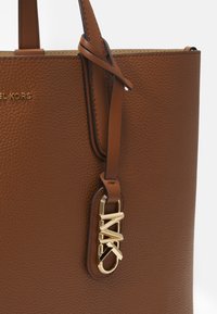 Hnědá taška přes rameno z strukturované kůže s zlatými kovovými detaily, která má výrazné logo a hladké kožené ucha. Jednoduchý, strukturovaný design.