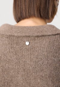 Pull en laine marron avec une finition douce et texturée et un motif subtil. Doté d'un bouton en argent rond au niveau de l'encolure arrière.