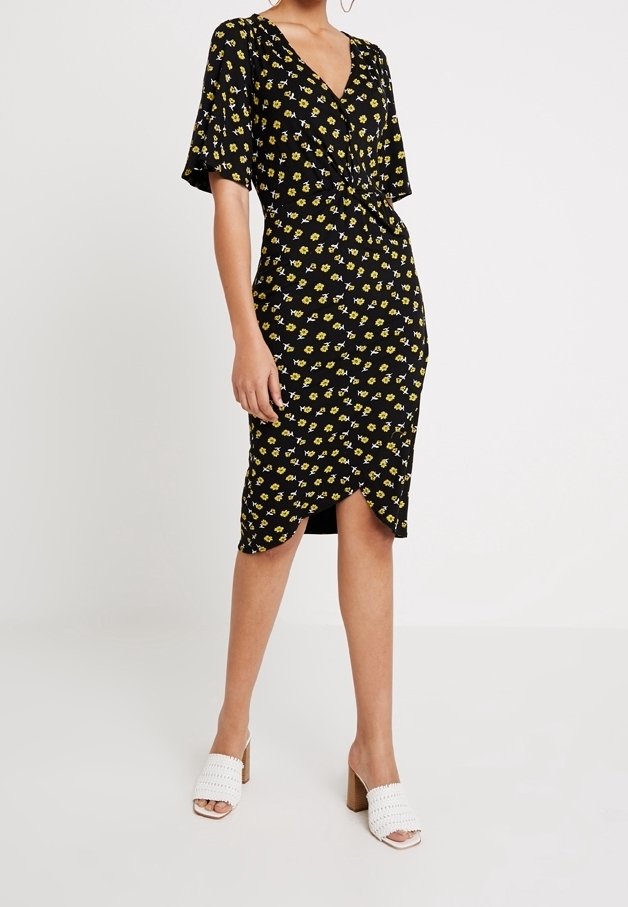 Femme portant une robe portefeuille midi noire avec imprimé floral jaune et manches courtes, associée à des sandales blanches à talons blocs ouverts.