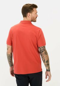 Rode polo shirt met een klassieke kraag en korte mouwen. Heeft een soepele textuur en zijslitsen. Model heeft zichtbare tatoeages op de arm.