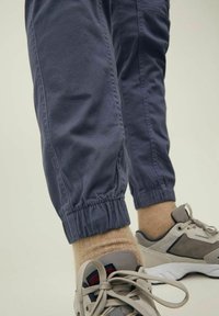 Detailní záběr na dolní části nohou, které mají na sobě námořnické tepláky s elastickými manžetami, béžové pono�žky a béžové šněrovací tenisky.