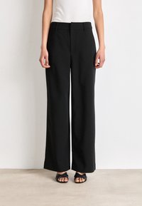 Svarta vida byxor i mjukt tyg med en figursydd midja och utan synliga fickor. Sammanfogade med svarta sandaler.