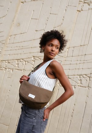Junge Frau in gestreiftem ärmellosem Oberteil und Jeans, die eine braune Umhängetasche trägt und gegen eine strukturierte beigefarbene Wand steht.