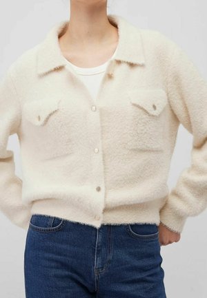 Kremowa, puszysta kurtka na guziki z kieszeniami na piersiach, noszona na białej bluzce i zestawiona z niebieskimi dżinsami.