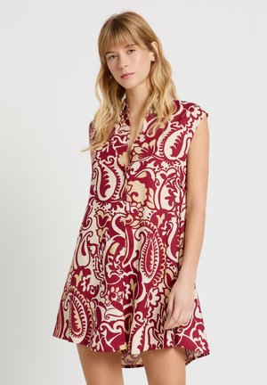 Jeune femme portant une robe sans manches rouge et beige à motif cachemire, debout devant un fond clair uni.