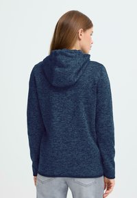 Sieviete ar gariem brūniem matiem, tērpusies tumši zilas kapucēta džemperī un gaiši pelēkos džinsos, stāv ar muguru pret vienkāršu baltu fona.