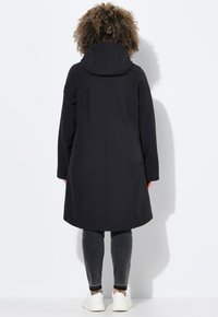 Manteau noir midi avec capuche, en tissu lisse. Il dispose de poches latérales et d'une coupe droite, associé à un jean foncé et des baskets blanches.