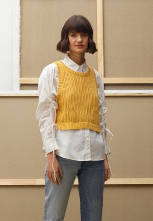 Maglione corto giallo lavorato a maglia con motivo a coste, indossato sopra una camicia bianca con colletto e maniche arricciate, abbinato a jeans blu strappati.