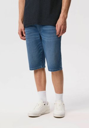 Dolna część ciała osoby noszącej niebieskie dżinsowe szorty do połowy uda, ciemną koszulę w prążki, białe skarpetki oraz białe trampki.