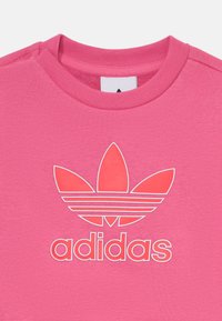 T-shirt en coton rose avec un logo Adidas blanc bien visible et trois accents en forme de feuilles, col ras du cou et tissu texturé.