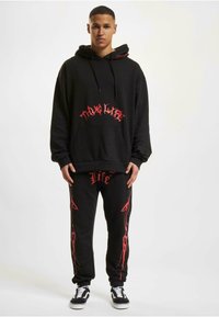 Svart hettegenser med rødt grafisk tekst og en frontlomme, kombinert med svarte bukser med røde detaljer og en logo, supplert med Vans-sko.