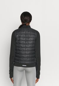 Chaleco acolchado negro con paneles cosidos, cuello alto y dobladillo acanalado. Lleva puesto sobre una blusa de manga larga, combinado con leggings grises.