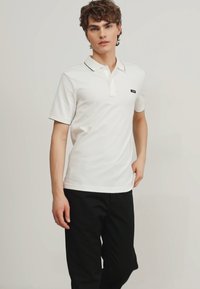 Polo bianco con colletto abbottonato, bordo nero sul colletto e un piccolo logo sul petto, abbinato a pantaloni neri.