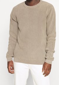 Pull en tricot beige avec texture côtelée, col rond et manches longues. Présente un ourlet et des poignets ajustés, associé à un pantalon blanc.