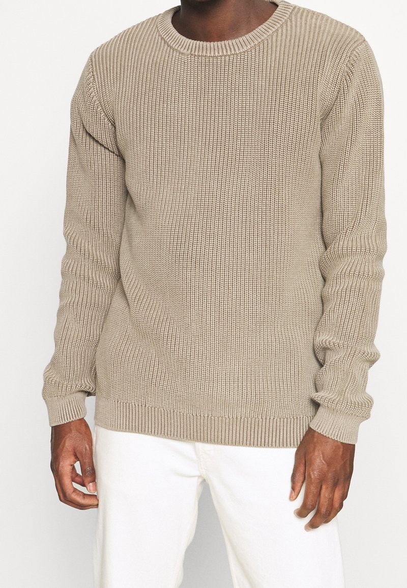 Pull en tricot beige avec texture côtelée, col rond et manches longues. Présente un ourlet et des poignets ajustés, associé à un pantalon blanc.