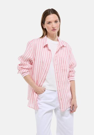 Femme aux cheveux bruns portant une chemise ample à rayures roses et blanches boutonnée, par-dessus un haut blanc et un pantalon blanc, debout avec une main dans la poche.
