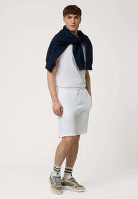 Shorts blancs à coupe décontractée avec cordon de serrage, associés à une chemise blanche à manches courtes et un sweat-shirt bleu marine drapé sur les épaules. Baskets beiges.