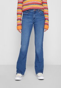 Blå, utsvingte jeans med midjehøyde, laget av denim. Har fem lommer og knappelukking. Kombinert med hvite sneakers.