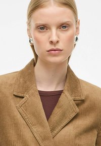 Blazer en velours côtelé marron avec un col cranté, porté sur un haut bordeaux. Tissu texturé avec des rayures verticales, agrémenté de boucles d'oreilles rondes en argent.