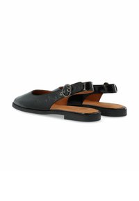 Fekete bőr slingback cipők lekerekített orral, lapos talppal és bokapánttal, amelyen egy kör alakú fém csat található. Texturált felület részlet.