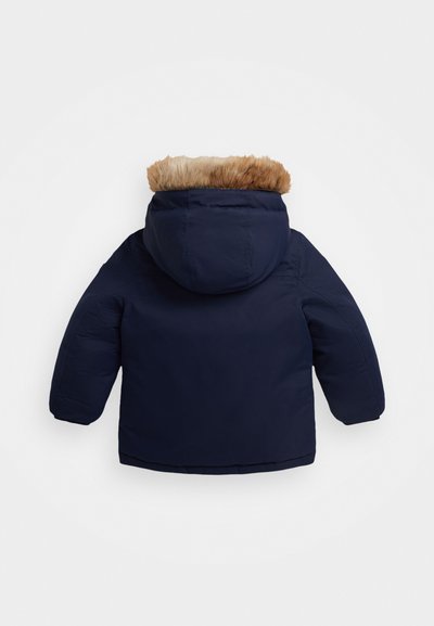 Veste matelassée bleu marine avec capuche en fourrure, présentant une texture lisse et des coutures piquées. Le manteau a des manches longues et une coupe mi-longue.