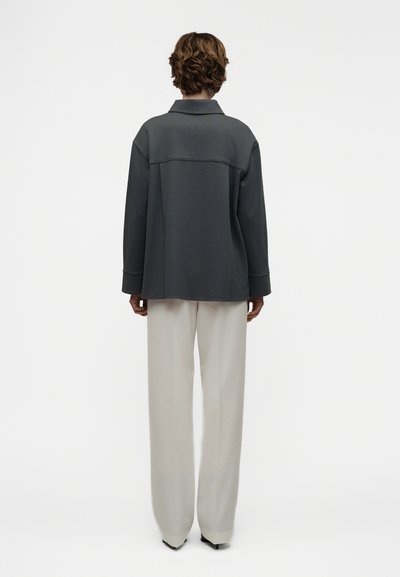 Άτομο με κοντά σγουρά μαλλιά στέκεται με την πλάτη προς τα εμπρός, φορώντας σκούρο γκρι φαρδιά ζακέτα και ανοιχτό μπεζ παντελόνι με φαρδιές γραμμές, σε απλό φόντο.