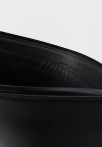 Wnętrze czarnej skórzanej torby o gładkiej strukturze, wyposażone w zamek błyskawiczny i wytłoczoną nazwę marki. Minimalistyczny design, brak dodatkowych detali.