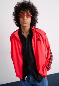 Red sports jacket with black accents and three stripes, zippered front, lightweight material, paired with a black button-up shirt and sunglasses.