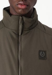 Olive green lightweight jacket with high collar, zip closure, and a textured fabric. Features a black logo patch on the left chest.