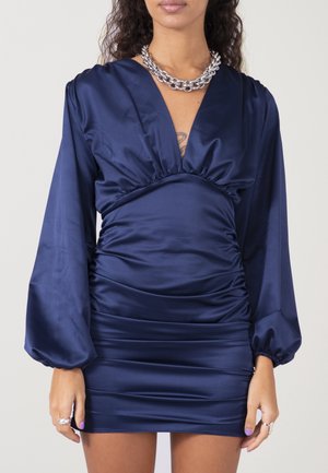 Žena v minišatech z tmavě modrého saténu s nabráním, dlouhými balónovými rukávy a stříbrným robustním řetízkovým náhrdelníkem, stojící před jednoduchým pozadím.