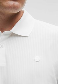 Biele polo tričko s textúrovaným pruhovaným dizajnom, dvoma gombíkmi pri golieri a subtílnym bielym logom na hrudi.