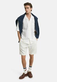 Weißes strukturiertes Kurzarmhemd, cremefarbene Shorts, dunkelblauer Strickpullover über den Schultern, braune Schuhe und weiße Socken mit blauen Streifen.