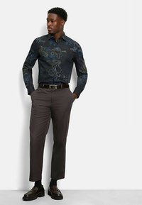 Langärmliges schwarzes Hemd mit blauem Blumenmuster, kombiniert mit grauen Hosen und dunklen Schuhen. Das Hemd hat einen Button-Down-Kragen.