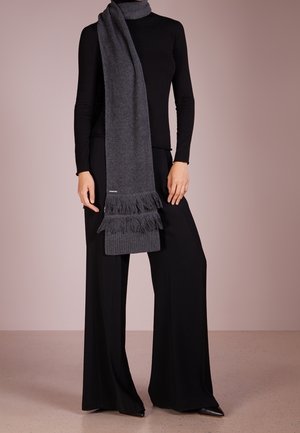 Personne portant un col roulé noir et un pantalon large, avec une écharpe grise foncée à franges drapée sur l'épaule, sur un fond uni.