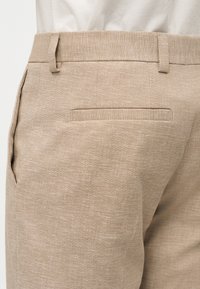 Pantalon en lin beige avec passants pour ceinture, poche latérale à la couture et poche passepoilée à l'arrière, porté avec une chemise blanche rentrée.