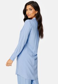 Jasnoniebieska bluzka w białe kropki, z długimi rękawami, zapinana na guziki przy mankietach oraz z okrągłym dołem. Wykonana z gładkiego, lekkiego materiału.