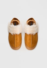 Un paio di pantofole in suede beige con morbido bordo in pelliccia bianca e fascia posteriore, presentate su uno sfondo chiaro e semplice.