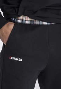 Crne trenirke s mekanom teksturom, sa karo pojasom i malim "KAVANAGH" logotipom u bijeloj i crvenoj boji na lijevom bedru.