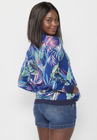 Giacca bomber blu con un vivace motivo di foglie tropicali nei colori rosa, verde e acquamarina. Polsi e orlo a costine accentuano il design. Abbinata a pantaloni shorts di denim.