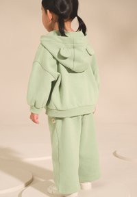 Grön bomullshoodie med öronformade detaljer på luvan, matchad med löst sittande byxor i samma färg. Vita sneakers fullbordar outfiten.