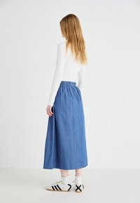 Gonna lunga in denim con vita elastica, tessuto blu, silhouette dritta, abbinata a sneakers bianche con strisce e dettagli neri.