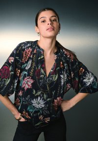 Blusa floreale in tessuto scuro con maniche a sbuffo, con fiori vivaci in rosso, rosa e verde. Design a scollo a V con scollo arricciato.