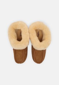 Chaussons en daim marron avec une doublure en laine beige, présentant une forme arrondie et une texture douce. Comprend des coutures latérales et une étiquette à l'extérieur.