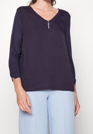 Femme portant un haut bleu marine foncé à manches longues avec une petite fermeture éclair devant et un pantalon large bleu clair, debout devant un fond uni.