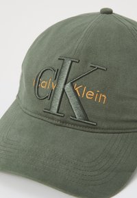 Groene katoenen pet met een gebogen rand, gekenmerkt door verhoogde donkergroene "CK" borduursel en oranje "Calvin Klein" tekst. Textuur van de stof is zichtbaar.