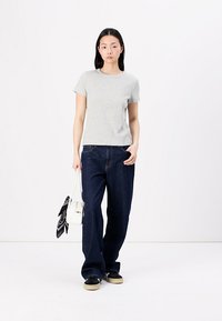 Grå t-shirt med korte ærmer, afslappet pasform, kombineret med mørkeblå brede bukser. Modellen holder en hvid hånd taske med et sort mønstret tørklæde.