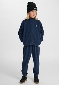Koordinerad marinblå fleece-träningsdräkt med zip-up jacka och elastiska byxor. Har detalj med liten logotyp och bärs med en svart mössa och sneakers.