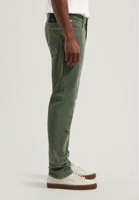 Pantalones de algodón verdes con un corte slim, que cuentan con bolsillos frontales y un parche con el logo en la parte trasera. Combinados con zapatillas blancas y una camiseta lisa.